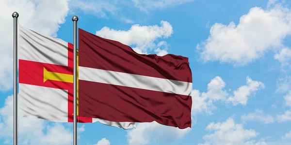 Guernsey e a bandeira da Letônia agitando no vento contra o céu azul nublado branco juntos. Conceito de diplomacia, relações internacionais . — Fotografia de Stock