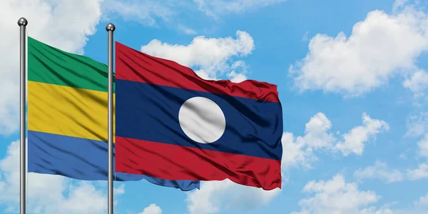 Gabón y la bandera de Laos ondeando en el viento contra el cielo azul nublado blanco juntos. Concepto diplomático, relaciones internacionales . — Foto de Stock