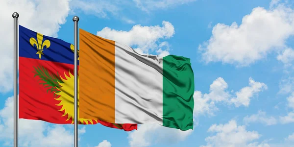 Guadalupe y la bandera de Costa de Marfil ondeando en el viento contra el cielo azul nublado blanco juntos. Concepto diplomático, relaciones internacionales . — Foto de Stock