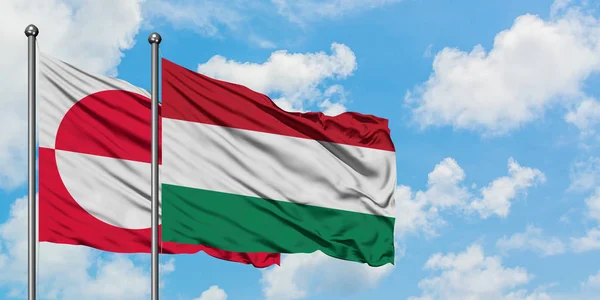 Bandera de Groenlandia y Hungría ondeando en el viento contra el cielo azul nublado blanco juntos. Concepto diplomático, relaciones internacionales . —  Fotos de Stock