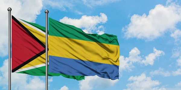 Bandera de Guyana y Gabón ondeando en el viento contra el cielo azul nublado blanco juntos. Concepto diplomático, relaciones internacionales . — Foto de Stock