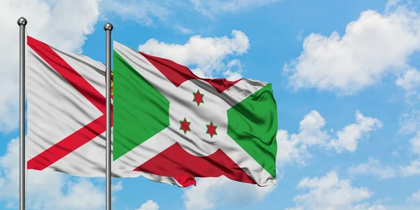 Bandera de Jersey y Burundi ondeando en el viento contra el cielo azul nublado blanco juntos. Concepto diplomático, relaciones internacionales . — Foto de Stock