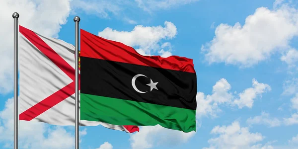 Bandera de Jersey y Libia ondeando en el viento contra el cielo azul nublado blanco juntos. Concepto diplomático, relaciones internacionales . — Foto de Stock