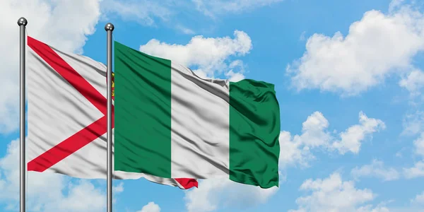 Bandera de Jersey y Nigeria ondeando en el viento contra el cielo azul nublado blanco juntos. Concepto diplomático, relaciones internacionales . — Foto de Stock