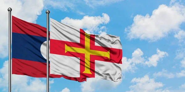 Laos y la bandera de Guernsey ondeando en el viento contra el cielo azul nublado blanco juntos. Concepto diplomático, relaciones internacionales . — Foto de Stock