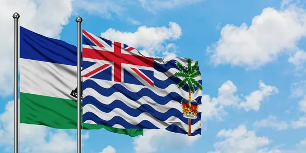 Bandera de Lesotho y del Territorio Británico del Océano Índico ondeando en el viento contra el cielo azul nublado blanco juntos. Concepto diplomático, relaciones internacionales . — Foto de Stock