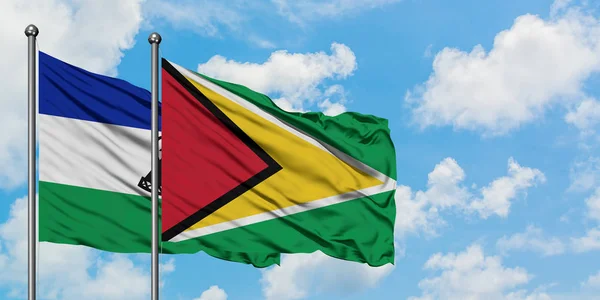 Lesoto e a bandeira da Guiana agitando no vento contra o céu azul nublado branco juntos. Conceito de diplomacia, relações internacionais . — Fotografia de Stock