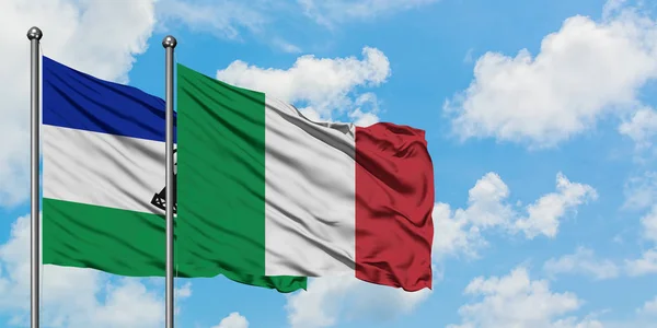 Lesotho y la bandera de Italia ondeando en el viento contra el cielo azul nublado blanco juntos. Concepto diplomático, relaciones internacionales . — Foto de Stock