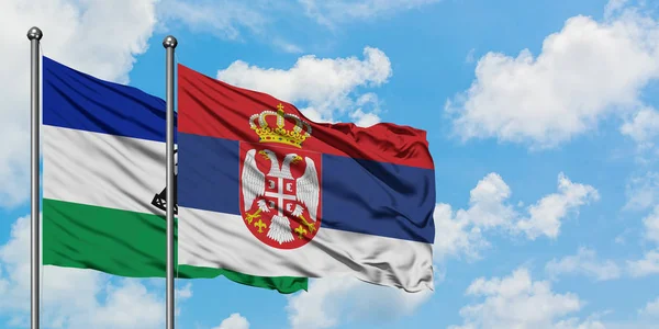 Lesotho y la bandera de Serbia ondeando en el viento contra el cielo azul nublado blanco juntos. Concepto diplomático, relaciones internacionales . — Foto de Stock
