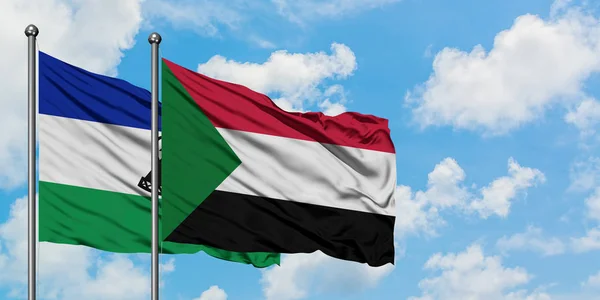 Lesotho y la bandera de Sudán ondeando en el viento contra el cielo azul nublado blanco juntos. Concepto diplomático, relaciones internacionales . — Foto de Stock
