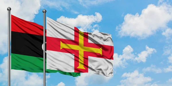 Bandera de Libia y Guernsey ondeando en el viento contra el cielo azul nublado blanco juntos. Concepto diplomático, relaciones internacionales . — Foto de Stock