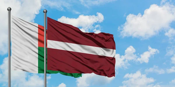 Drapeau de Madagascar et de Lettonie agitant dans le vent contre ciel bleu nuageux blanc ensemble. Concept de diplomatie, relations internationales . — Photo
