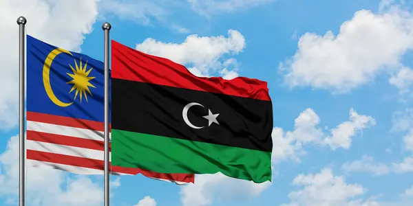 Bandera de Malasia y Libia ondeando en el viento contra el cielo azul nublado blanco juntos. Concepto diplomático, relaciones internacionales . — Foto de Stock