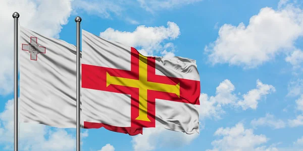 Malta e la bandiera di Guernsey sventolano insieme nel vento contro il bianco cielo blu nuvoloso. Concetto di diplomazia, relazioni internazionali . — Foto Stock