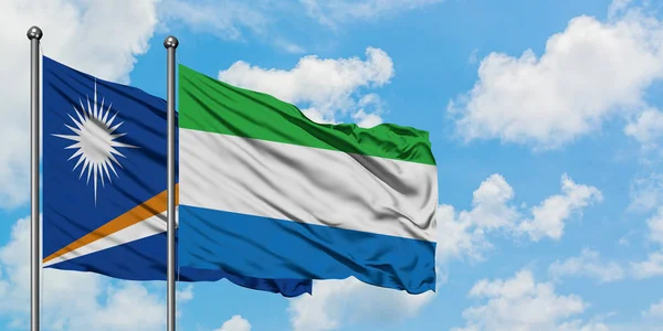 Marshallův ostrov a vlajka Sierra Leone mávali ve větru proti bíle zatažené modré obloze. Diplomacie, mezinárodní vztahy. — Stock fotografie