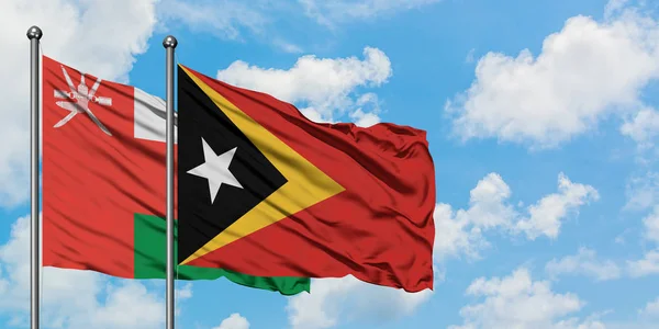 Omán y Timor Oriental ondean en el viento contra el cielo azul nublado blanco juntos. Concepto diplomático, relaciones internacionales . — Foto de Stock