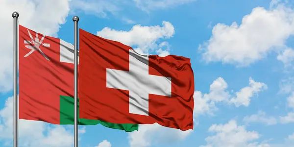 Omán a Švýcarsko mávali ve větru proti bíle zatažené modré obloze. Diplomacie, mezinárodní vztahy. — Stock fotografie