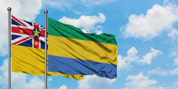 Niue i Gabon flaga machając w wiatr przed białym zachmurzone błękitne niebo razem. Koncepcja dyplomacji, stosunki międzynarodowe. — Zdjęcie stockowe