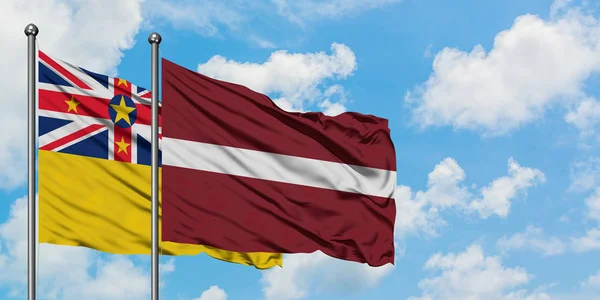 Niue e a bandeira da Letônia agitando no vento contra o céu azul nublado branco juntos. Conceito de diplomacia, relações internacionais . — Fotografia de Stock