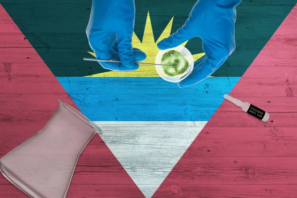 Antigua and Barbuda flag on laboratory table. Medical healthcare technologist holding COVID-19 swab collection kit, wearing blue protective gloves, epidemic concept.
