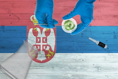 Laboratuvar masasında Sırbistan bayrağı var. Sağlık teknolojisi uzmanı COVID-19 bez toplama kiti tutuyor, mavi koruyucu eldivenler giyiyor, salgın konsepti.