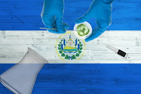 El Salvador flag on laboratory table. Medical healthcare technologist holding COVID-19 swab collection kit, wearing blue protective gloves, epidemic concept.