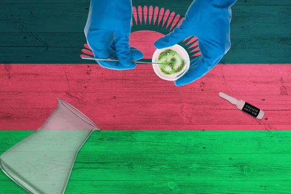 Malawi flag on laboratory table. Medical healthcare technologist holding COVID-19 swab collection kit, wearing blue protective gloves, epidemic concept.