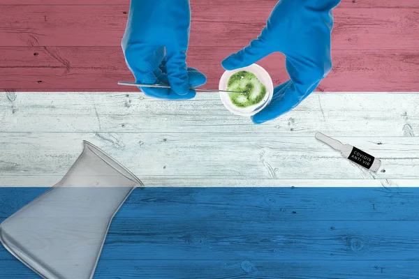 Netherlands flag on laboratory table. Medical healthcare technologist holding COVID-19 swab collection kit, wearing blue protective gloves, epidemic concept.