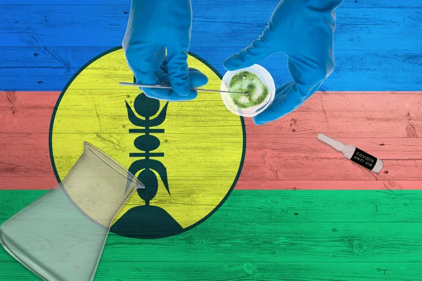 New Caledonia flag on laboratory table. Medical healthcare technologist holding COVID-19 swab collection kit, wearing blue protective gloves, epidemic concept.