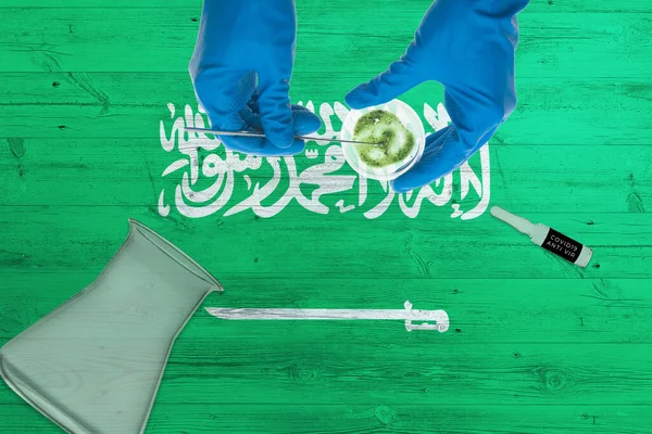 Saudi Arabia flag on laboratory table. Medical healthcare technologist holding COVID-19 swab collection kit, wearing blue protective gloves, epidemic concept.