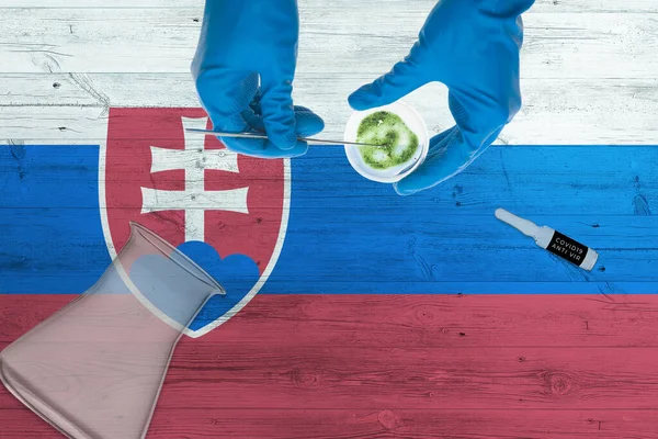 Slovakia flag on laboratory table. Medical healthcare technologist holding COVID-19 swab collection kit, wearing blue protective gloves, epidemic concept.