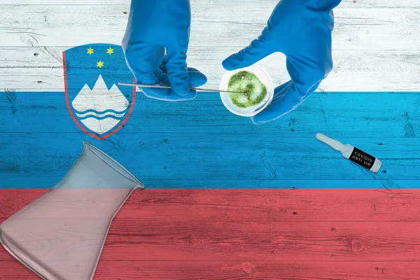 Slovenia flag on laboratory table. Medical healthcare technologist holding COVID-19 swab collection kit, wearing blue protective gloves, epidemic concept.