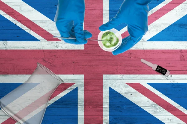 United Kingdom flag on laboratory table. Medical healthcare technologist holding COVID-19 swab collection kit, wearing blue protective gloves, epidemic concept.