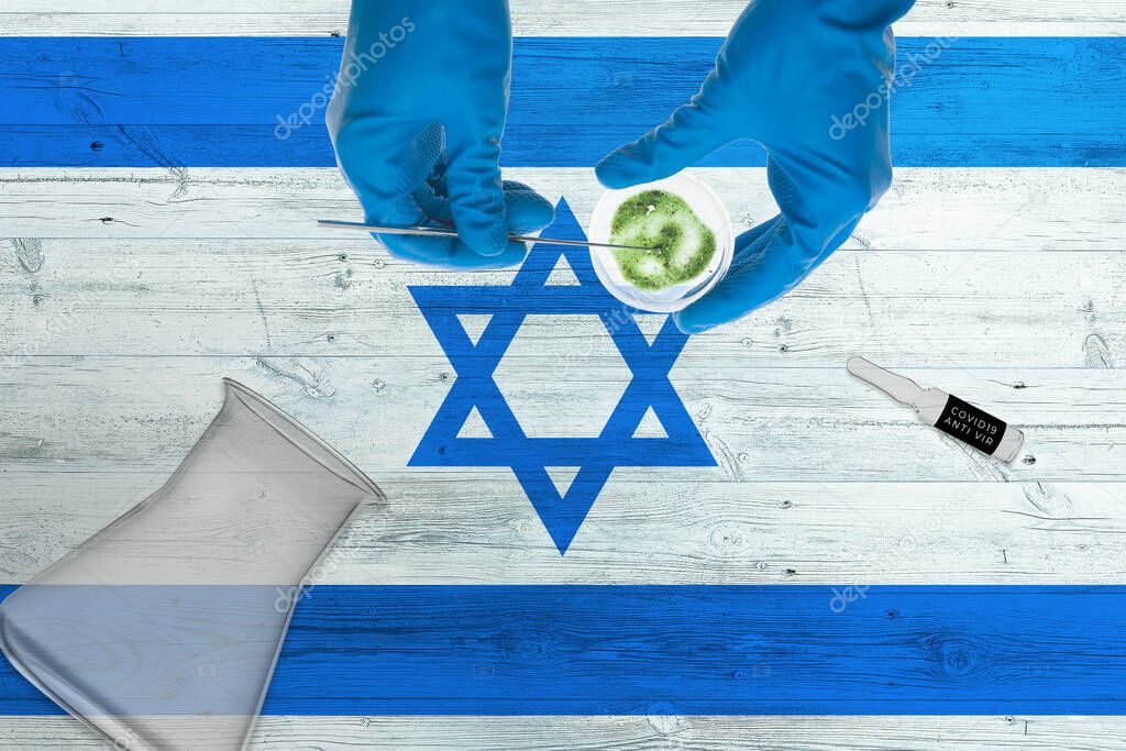 Israel flag on laboratory table. Medical healthcare technologist holding COVID-19 swab collection kit, wearing blue protective gloves, epidemic concept.