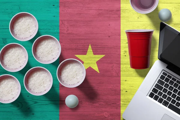 Cameroon flag concept with plastic beer pong cups and laptop on national wooden table, top view. Beer Pong game.