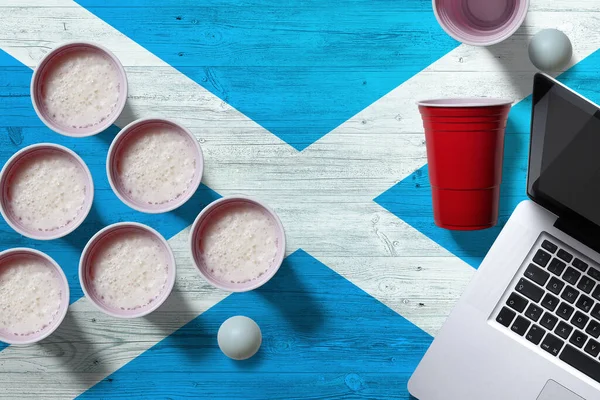 Scotland flag concept with plastic beer pong cups and laptop on national wooden table, top view. Beer Pong game.