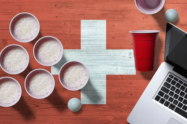 Switzerland flag concept with plastic beer pong cups and laptop on national wooden table, top view. Beer Pong game.