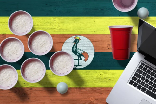 Uganda flag concept with plastic beer pong cups and laptop on national wooden table, top view. Beer Pong game.