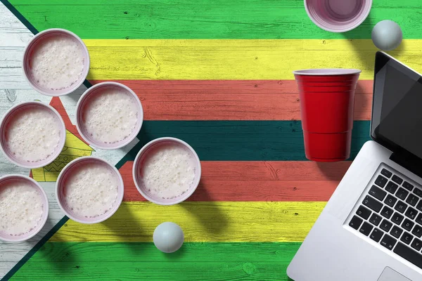 Zimbabwe flag concept with plastic beer pong cups and laptop on national wooden table, top view. Beer Pong game.