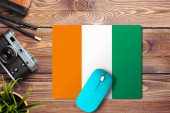 Pobřeží slonoviny vlajka na dřevěném pozadí s modrou bezdrátovou myší na podložce myši, horní pohled. Koncept digitálních médií.