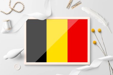 Beyaz yaratıcı arka planda ahşap çerçeveli Belçika bayrağı. Beyaz tema, tüy, papatya, düğme, kurdele nesneleri.