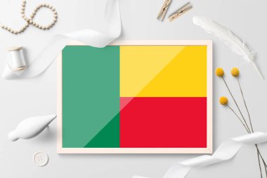 Beyaz yaratıcı arka planda ahşap çerçeveli Benin bayrağı. Beyaz tema, tüy, papatya, düğme, kurdele nesneleri.