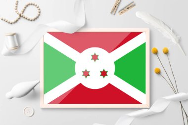 Beyaz yaratıcı arka planda ahşap çerçeveli Burundi bayrağı. Beyaz tema, tüy, papatya, düğme, kurdele nesneleri.