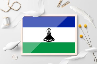Beyaz yaratıcı arka planda ahşap çerçeveli Lesotho bayrağı. Beyaz tema, tüy, papatya, düğme, kurdele nesneleri.