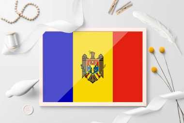 Beyaz yaratıcı arka planda tahta çerçeveli Moldova bayrağı. Beyaz tema, tüy, papatya, düğme, kurdele nesneleri.