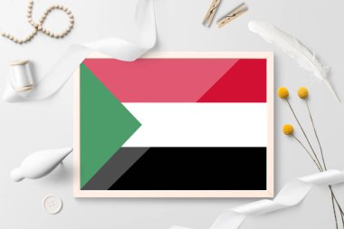 Beyaz yaratıcı arka planda tahta çerçeveli Sudan bayrağı. Beyaz tema, tüy, papatya, düğme, kurdele nesneleri.