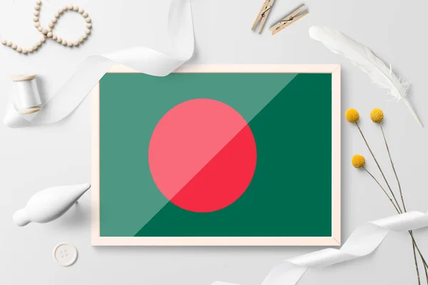 Bangladesh flag in wooden frame on white creative background. White theme, feather, daisy, button, ribbon objects.