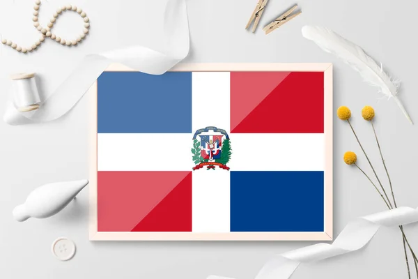 Dominican Republic flag in wooden frame on white creative background. White theme, feather, daisy, button, ribbon objects.