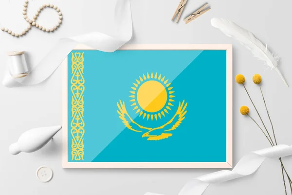 Kazakhstan flag in wooden frame on white creative background. White theme, feather, daisy, button, ribbon objects.