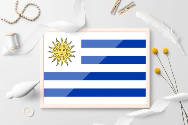 Uruguay flag in wooden frame on white creative background. White theme, feather, daisy, button, ribbon objects.